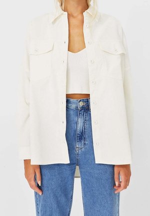 Woman wearing an open off-white button-up shirt over a white cropped knit top paired with high-waisted blue jeans.