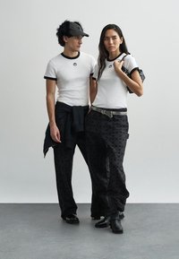 Bela rebrasta majica s črnimi poudarki, nošena s črnimi hlačami z vzorci. Dodatki vključujejo črno kapo in pas. Minimalističen dizajn.