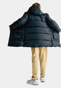 Donkerblauwe pufferjas met een capuchon, voorzien van gequiltte delen en zijzakken. Draag je over lichtbeige broek en grijze sneakers.