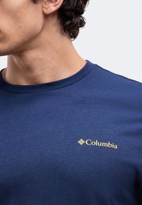 Άνδρας που φορά μπλε μπλουζάκι Columbia με μικρό κίτρινο λογότυπο Columbia στο αριστερό στήθος, κοιτάζει αριστερά με το κεφάλι μερικώς ορατό.