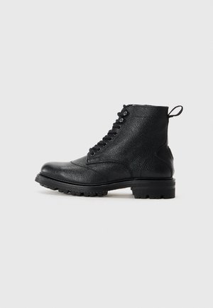 Botas de tobillo de cuero negro con una superficie texturizada, puntera redonda, cordones en la parte frontal y suela rugosa, con una lengüeta en la parte posterior.