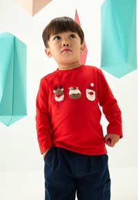 Chemise rouge à manches longues avec trois motifs de Noël brodés : un renne, un pudding et le Père Noël. Un pantalon bleu marine complète la tenue.