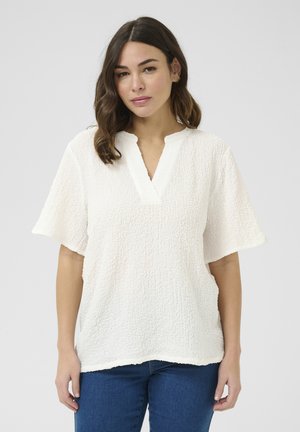 Blouse blanche texturée à manches courtes avec un petit col en V. Présente une coupe fluide et un ourlet brut. Portée avec un jean en denim bleu.