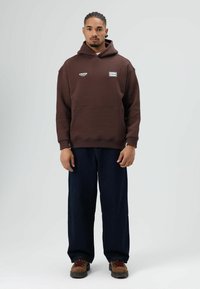 Bruine hoodie met voorzak, geborduurde logo's en loszittende donkere spijkerbroek. Draagbaar met bruine laarzen met rode veters.