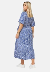 Sininen maximekko, jossa on valkoinen abstrakti kuvio, lyhyet flare-hihat ja solmittava vyötärö, yhdistettynä valkoisiin tennareihin rennon ilmeen saavuttamiseksi.