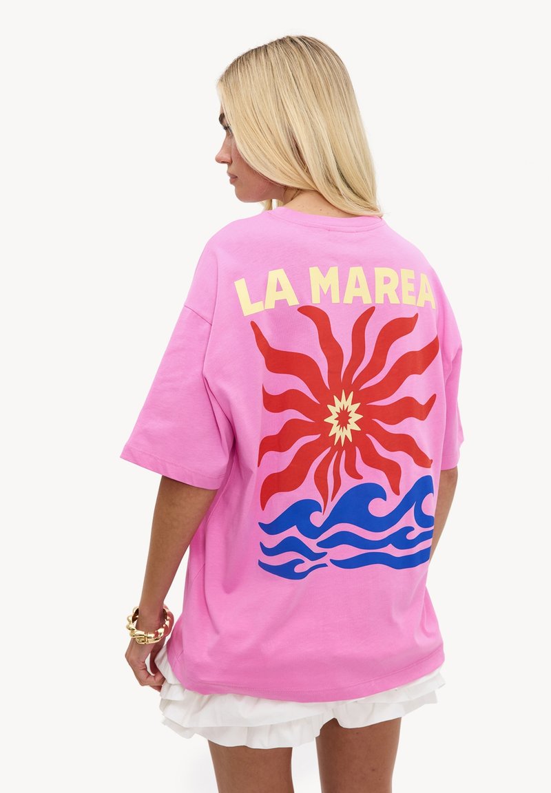 Femme blonde de profil portant un t-shirt rose avec le texte "LA MAREA", un éclat de soleil rouge et des graphismes de vagues bleues dans le dos ainsi qu'une jupe blanche à volants.