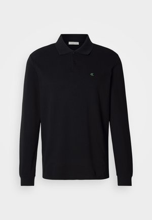 Černé polo tričko s dlouhým rukávem vyrobené z bavlny, s tříknoflíkovou légou a malým zeleným vyšitým logem na levé straně hrudi.