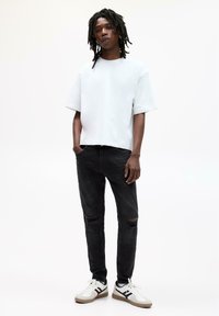 Ung mand med dreadlocks står iført en hvid oversized t-shirt, slidte sorte jeans og hvide sneakers foran en hvid baggrund.