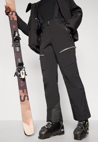 Schwarze Skihosen mit Reißverschluss-Taschen, stabilen Riemen und einer glatten Textur. Ein Paar Skier und Skischuhe werden in der Hand gehalten, wobei die Bindungen zu sehen sind.