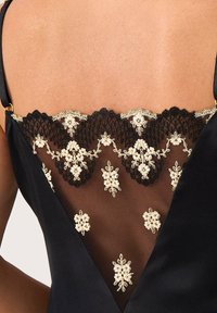 Czarna odzież z tyłem w kształcie litery V z misternym złotym i kościanym haftem kwiatowym na przezroczystej czarnej koronce. Gładki materiał, regulowane ramiączka.