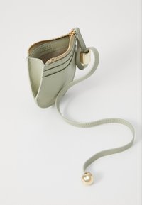 Borsa in pelle verde con dettagli in oro, chiusura con zip e tracolla staccabile con un elemento decorativo a forma di pallina. Design compatto ideale per i necessari.