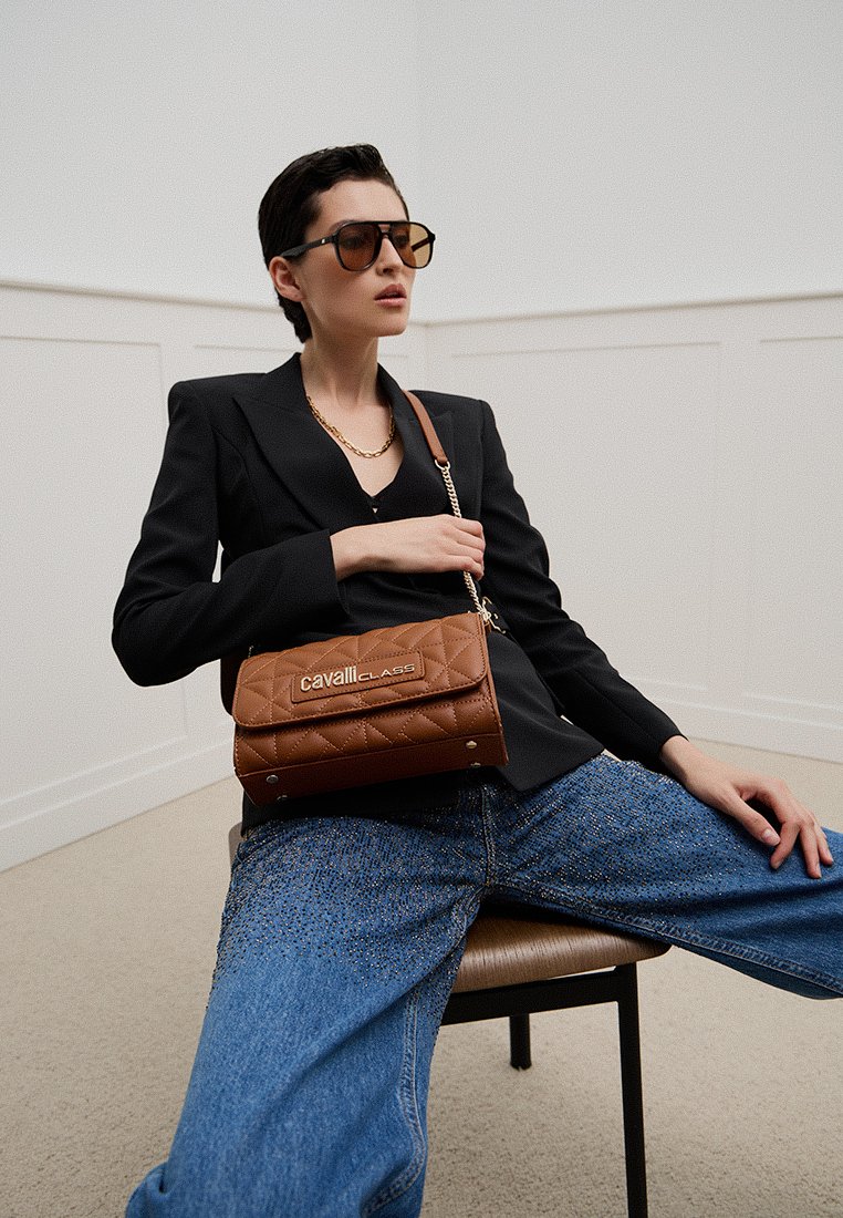 Sac à main marron matelassé avec rabat et détail de logo, équipé d'une bandoulière en chaîne. Le modèle porte un blazer noir et un jean bleu clair.