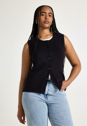 Ung kvinde med lange fletninger iført en sort knappet ærmeløs vest over en hvid top og lyseblå jeans mod en ensfarvet baggrund.