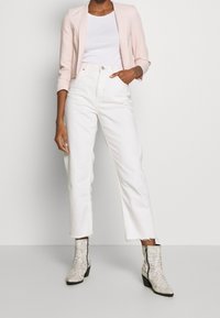 Persoon draagt een lichtroze cropped blazer, witte top, hooggetailleerde witte jeans met rafelige zoom en witte enkellaarsjes met slangenprint.