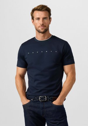 Man met kort bruin haar die een donkerblauw Hackett T-shirt en donkere spijkerbroek draagt met zijn handen in zijn zakken tegen een effen achtergrond.