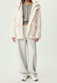 Roze, donzige jas met rits en capuchon, grijs overhemd met knopen en losse grijze joggingbroek. Witte sneakers maken de outfit compleet.