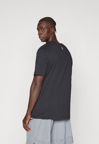 Musta puuvillainen lyhythihainen T-paita, jossa on pyöreä kaula-aukko ja pieni keltainen logo takana. Yhdistetty harmaisiin shortseihin. Yksinkertainen, rento istuvuus.