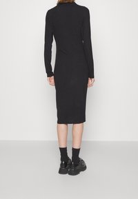 Schwarzes geripptes Langarmkleid mit taillierter Passform, knielang und subtiler Textur. Getragen mit schwarzen Knöchelsocken und Schnürschuhen.