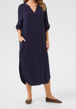 Woman wearing a loose-fitting, long navy dress with rolled-up sleeves and side pockets, paired with brown flat shoes.