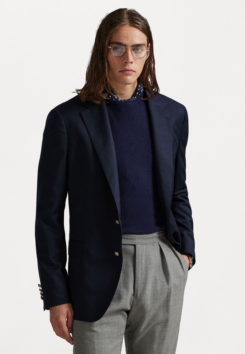 Blazer azul-marinho com botões dourados, usado sobre um suéter de malha azul-escuro e calças plissadas cinza claras, destacando-se uma camisa de bolinhas por baixo.