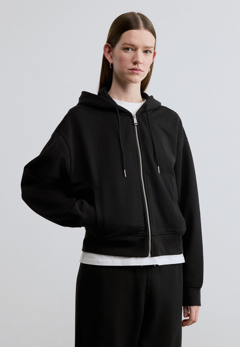 Μαύρο φούτερ με φερμουάρ και άνετη γραμμή, τσέπες καγκουρό, κουκούλα με κορδόνι και ριμπ μανσέτες. Φορώντας πάνω από λευκό μπλουζάκι και μαύρο παντελόνι.