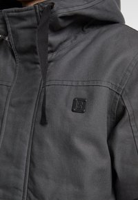 Juodas gobtuvu aprengtas švarkas, pagamintas iš tekstūruoto audinio, su siūlėmis, priekinėmis kišenėmis ir kvadratine logotipo etikete ant krūtinės.