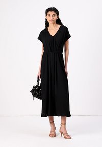 Black, short-sleeve maxi dress in a smooth fabric, featuring a V-neck and a tied waist, paired with strappy heels and a textured handbag.