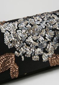 Svart tygclutch prydd med stora metalliska silver- och bronseconominer i ett blommönster. Slät textur med glänsande detaljer.