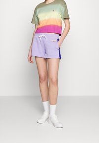 En person som bär en kortärmad tröja med grön till lila gradient och lavendel färgade shorts med dragsko, tillsammans med vita sneakers och strumpor, står mot en enfärgad bakgrund.