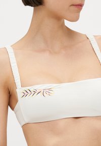 Balts bandeau bikini tops, izgatavots no gludas materiāla, ar krāsainiem ziedu izšuvumiem kreisajā pusē un savītiem siksniņām.