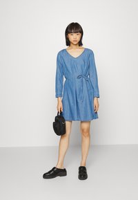 Vestido de ganga com mangas longas, decote em V, cintura com cordão e saia rodada. Usado com sapatos pretos chunky de slip-on e uma pequena bolsa preta.
