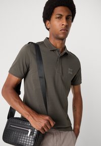 Polo de culoare verde măsliniu închis cu un guler ribbed și un patch mic cu logo, asortat cu o geantă crossbody texturată de culoare neagră, dotată cu un fermoar argintiu.