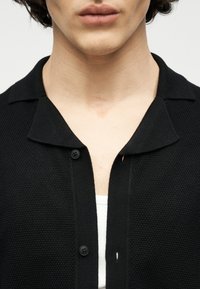 Prim-plan cu o persoană purtând un cardigan texturat negru peste un tricou alb, arătând gulerul și o parte a pieptului.