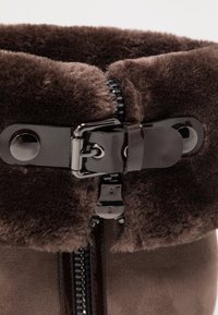 Brązowe botki z imitacji futra, wyłożone miękkim materiałem, mają połyskujący czarny zamek błyskawiczny oraz klamrę.