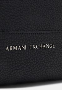 Sort lædertaske med en tekstureret overflade. Inkluderer et sølvfarvet "Armani Exchange" logo på fronten, fremhævet af et rent, minimalistisk design.