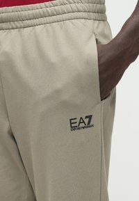 Bézs színű melegítőnadrág rugalmas derékpánttal, oldalzsebbel és fekete EA7 Emporio Armani logóval a combon. Simább anyag textúra.