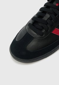 Black sneaker featuring suede and leather materials, a lace-up design, red accent stripes, and a textured rubber sole with a geometric pattern.