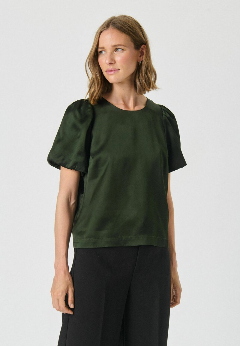 Top in satin verde scuro con maniche a sbuffo corte e scollatura rotonda, caratterizzato da una texture liscia e una vestibilità rilassata. Abbinato a pantaloni neri.