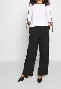 Blouse blanche avec des bords noirs et des liens sur les côtés, associée à un pantalon noir taille haute à jambes larges et des chaussures à talons noires. Tissus lisses et coupe moderne.