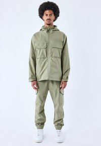 Olijfgroene, lichte, gestructureerde jas met een rits aan de voorkant en meerdere zakken, gecombineerd met bijpassende elastische broek en witte sneakers.