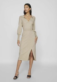 Beige gebreid jurk met een kraagvormige V-hals, lange pofmouwen, getailleerde taille en een zijsplit. Glad textuur met een lengte tot halverwege de kuit.