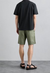 Homme debout face au mur portant un t-shirt noir, un short olive et des sandales à boucle noires sur un sol gris.