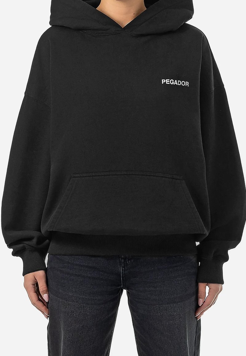 Personne portant un sweat à capuche noir avec poche avant et texte blanc "PEGADOR" sur la poitrine, associé à un jean foncé.
