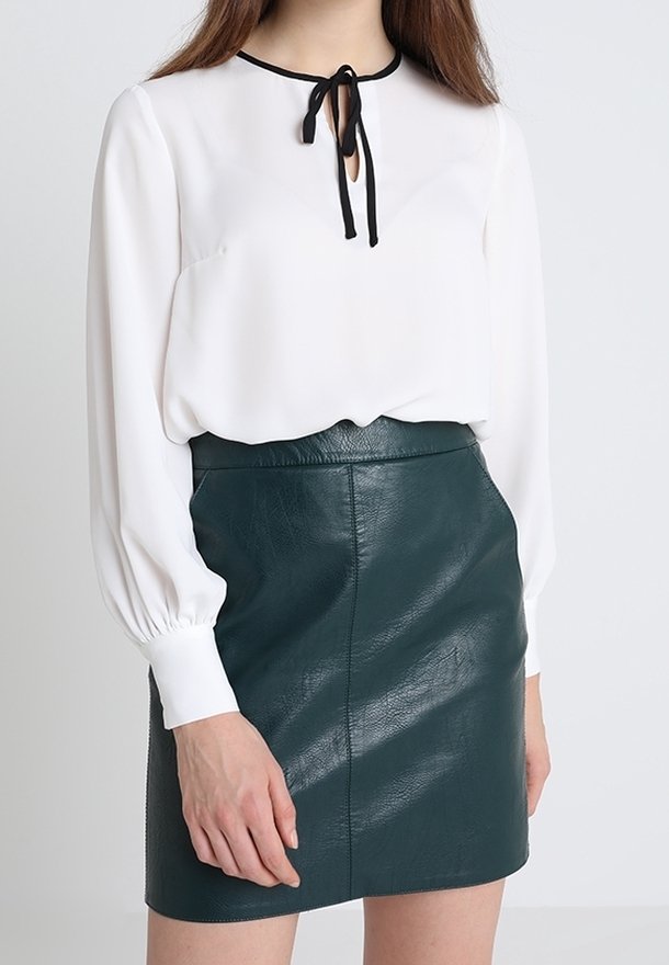 Femme portant un chemisier blanc à manches longues avec un détail de cravate noire et une mini-jupe en cuir vert foncé.
