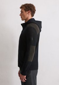 Junger Mann im Profil, der einen schwarzen Strick-Zip-Hoodie mit beigen Patches an Ärmeln und Schultern trägt, steht vor einer weißen strukturierten Wand.