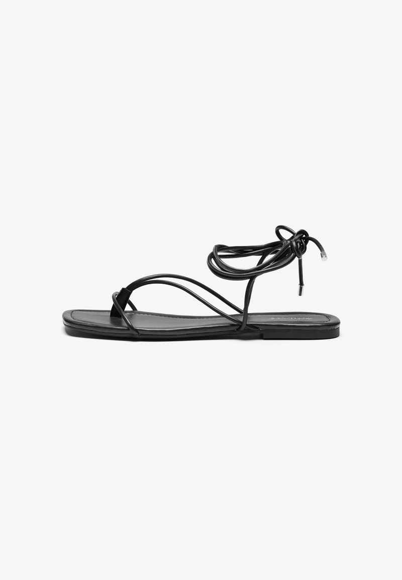 Sorte flate sandaler med tynne, fleksible stropper, som har et knyttet design ved ankelen, og en glatt, rektangulær fotbed.