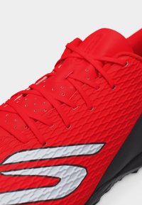Κόκκινο αθλητικό παπούτσι με υφή στην επιφάνεια, κόκκινες κορδόνια και μεγάλο λευκό στυλιζαρισμένο λογότυπο "S" στο πλάι, τοποθετημένο σε μαύρη σόλα.