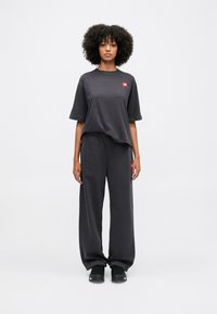 T-shirt nera oversized a maniche corte con un piccolo logo rosso, abbinata a pantaloni tuta neri larghi. Scarpe nere completano l'outfit.