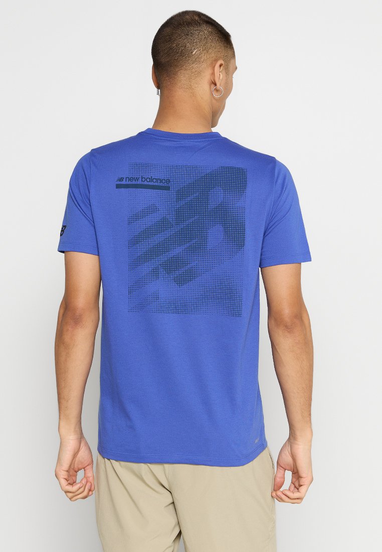 Hombre vistiendo una camiseta azul de New Balance con un diseño de logo pixelado negro en la parte posterior, combinada con pantalones cortos beige, de pie contra un fondo blanco.