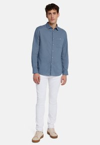 Camicetta azzurro chiaro con maniche lunghe, tasca sul petto e vestibilità morbida, abbinata a jeans bianchi e sneakers beige.